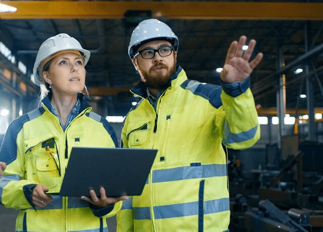 Two people in yellow jackets and hard hats holding a laptop.